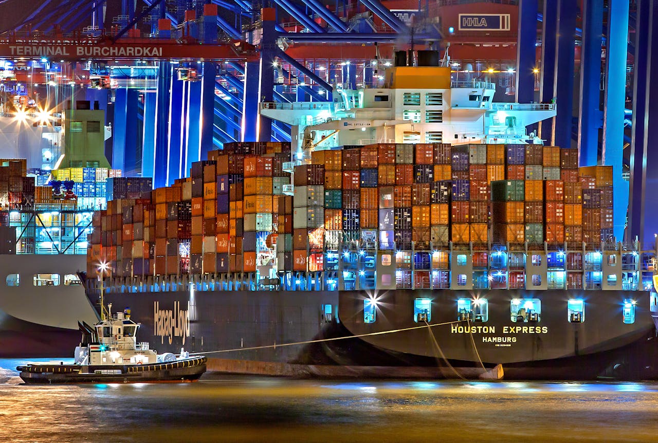 why-choose-us A brightly lit cargo ship at Hamburg harbor with stacked containers and a tugboat.